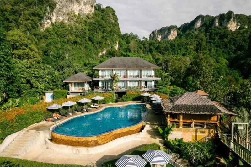 Phutawan Resort Krabi features a central outdoor pool, sun loungers, umbrellas, a multi-story building, and lush green mountains in the background for a perfect Krabi resort escape.