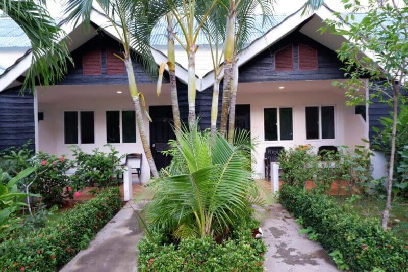 Two adjacent bungalow-style units with black and white exteriors, surrounded by greenery and palm trees, create a serene Krabi Homestay vibe, with a walkway leading to the doors.
