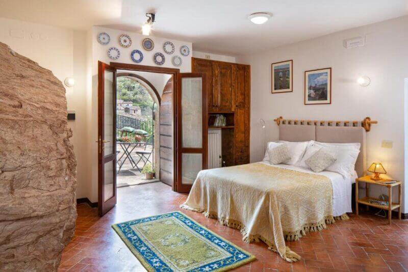 Bedroom with a double bed, wooden furniture, a rock wall feature, wall-mounted plates, and glass doors opening to an outdoor patio with table and chairs at Residence Menotre in the charming village of Rasiglia.