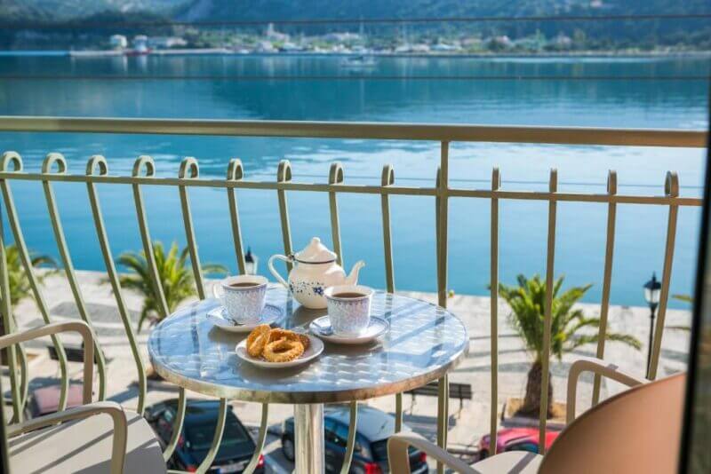 A round table at a Tourist Boutique Hotel balcony in Argostoli overlooks a calm waterfront. The table has two cups of coffee, a teapot, and pastries. Palm trees and parked cars are visible below.