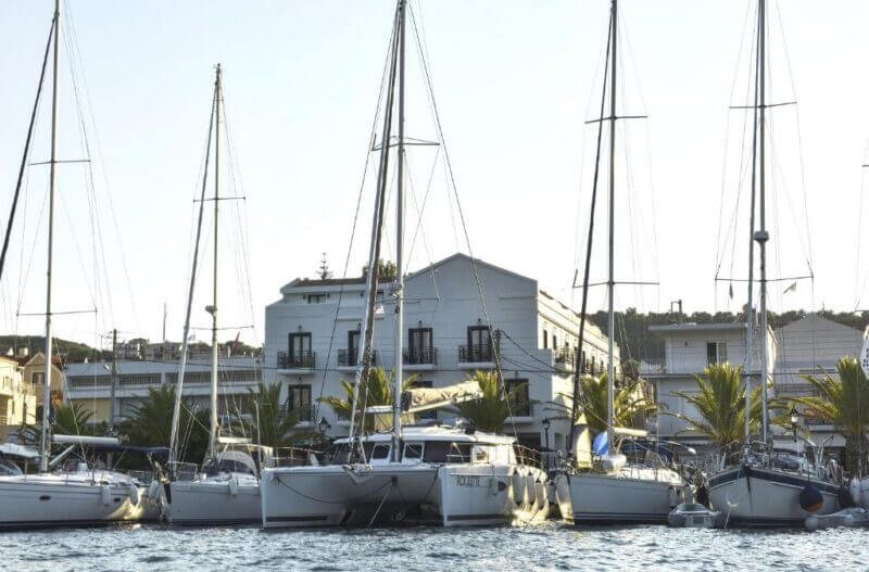 Several sailboats are docked in a marina in front of a grand white two-story building with balconies and palm trees, under the clear sky of Argostoli, Kefalonia.