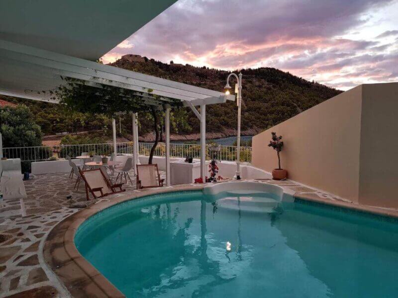 Outdoor pool with patio seating, white pergola, potted plants, and a view of hills and a colorful sunset sky—an idyllic Vassilis Retreat setting for relaxation.