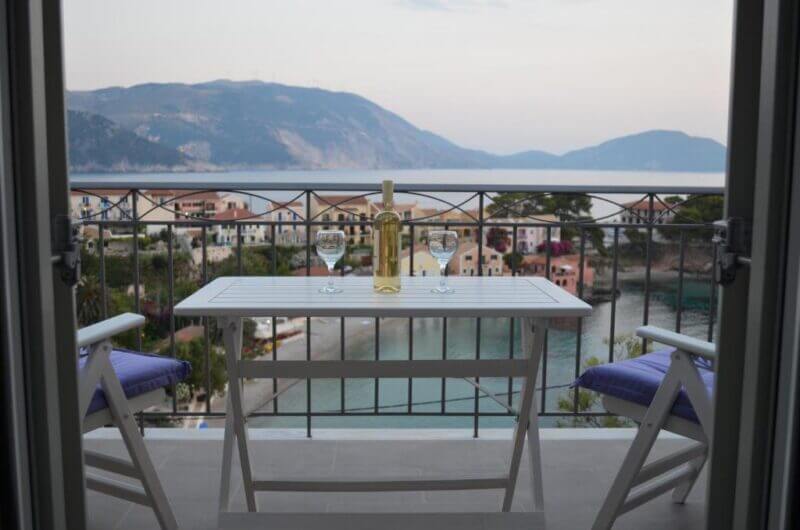 A balcony at Romanza Studios is set for two with wine and glasses, overlooking the charming coastal village of Asos, the sea, and distant mountains.