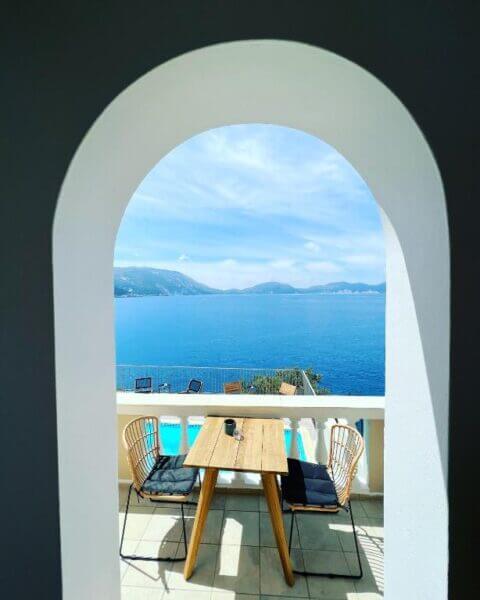 View through an arched doorway of a balcony at Asos Kanakis Apartments, featuring a wooden table, two chairs, and a scenic backdrop of blue sea and mountains under a clear sky.