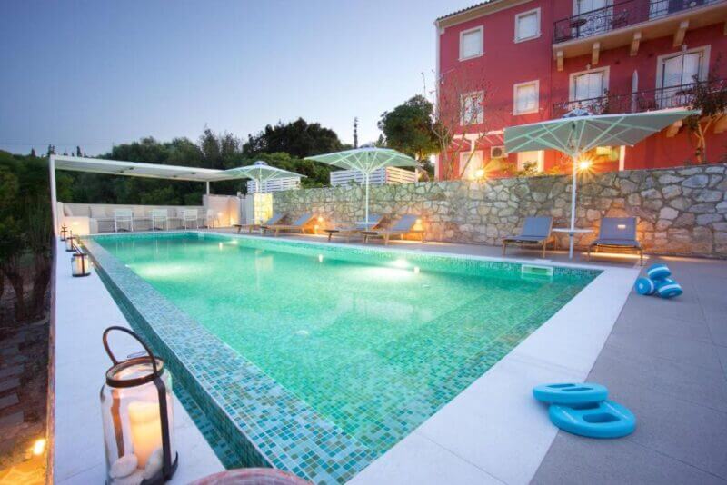 Rectangular outdoor swimming pool with mosaic tiles at Fiskardo Apartments, featuring poolside loungers, large umbrellas, and soft evening lighting beside a stone wall and a red building.