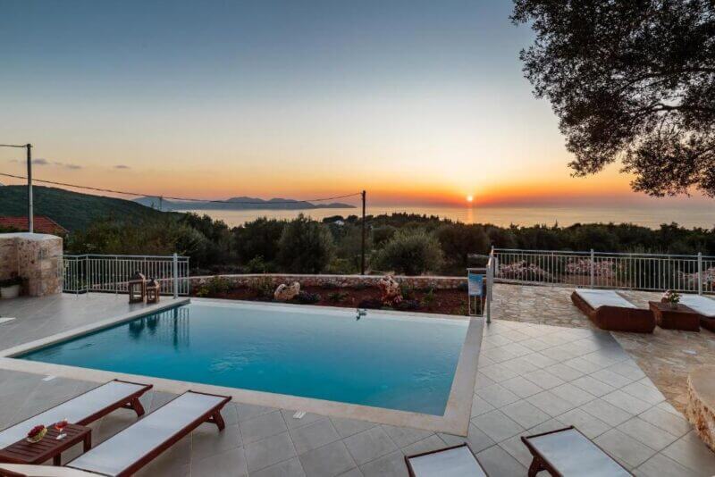 Rectangular outdoor pool with lounge chairs on a tiled patio at Fiskardo Villa overlooks trees and the sea at sunset, with a clear sky and distant hills in the background.