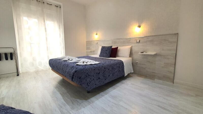 Modern bedroom at Hostal Met Madrid with a double bed in blue bedding, wall lamps above the headboard, white walls, wood-look flooring, and natural light from a window—ideal for those seeking comfortable budget accommodation in Madrid.