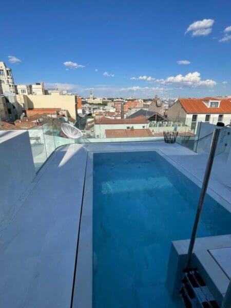 Rooftop pool with clear glass fencing at 60 Balconies Madrid overlooks a cityscape with tiled rooftops and buildings under a blue sky with scattered clouds.