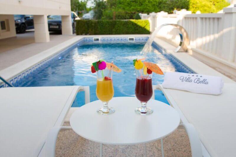 Two colorful cocktails sit on a small table between lounge chairs beside a sparkling pool, with a towel labeled Villa Bella in the background—an inviting scene at Villa Apartments near Art Trogir.