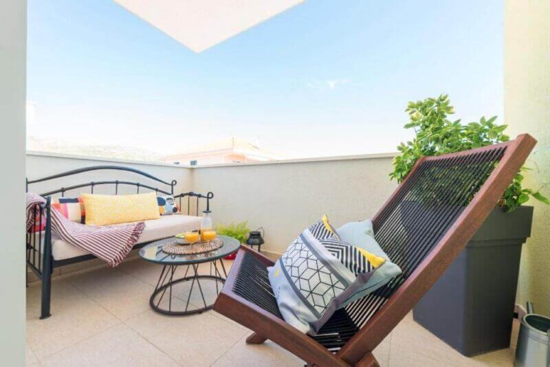 A small Narancin Trogir balcony with a lounge chair, daybed, round glass table with drinks, and potted plants under a clear sky.