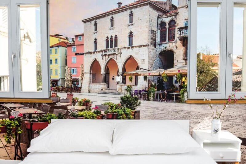 A bed with white pillows and linens is positioned in front of a wall mural depicting a European street scene, reminiscent of Villa Galla Split, with historic buildings, arches, and charming outdoor cafes.