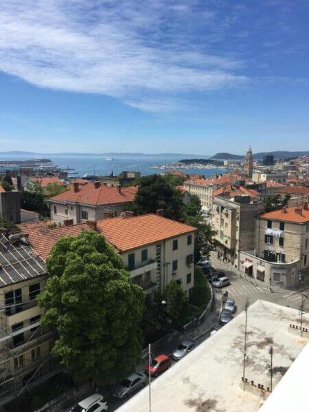 Enjoy a stunning view from Apartment Marija in Split, overlooking red-roofed buildings, a tree-lined street, and the sparkling sea under a partly cloudy sky.