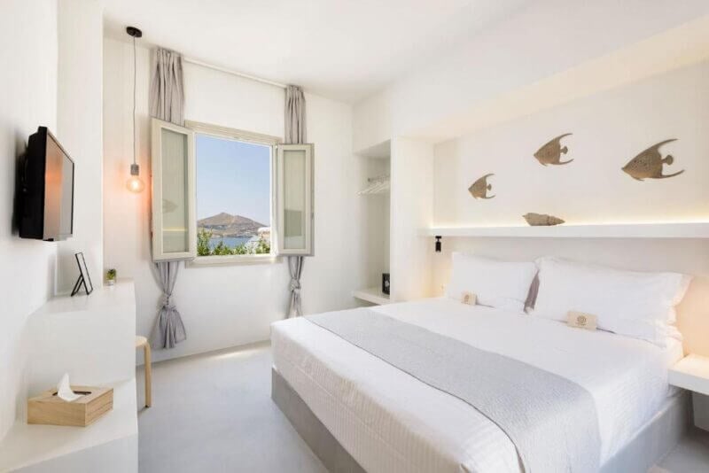 Bright, minimalist bedroom at Peven Suites Paros with a double bed, light gray linens, open window offering a scenic view, wall-mounted TV, and fish decorations above the bed.