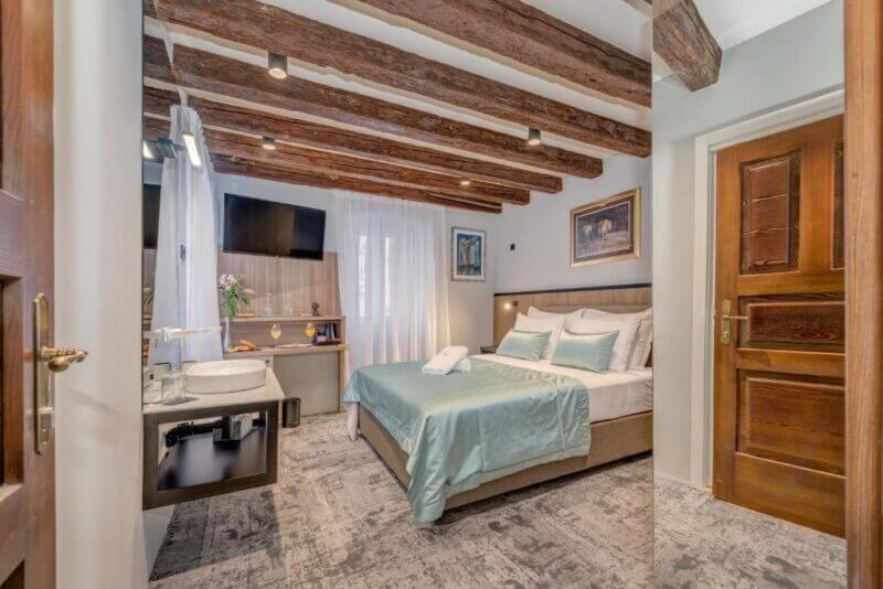 A cozy bedroom at Makarun Heritage Rooms in Split, featuring exposed wooden ceiling beams, a double bed with teal bedding, a wall-mounted TV, a sink, and a large window with sheer curtains.