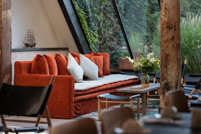 A modern lounge area at Hôtel Saint Nicolas features an orange sofa, light cushions, a wooden coffee table with a vase of flowers, and large windows overlooking lush greenery.