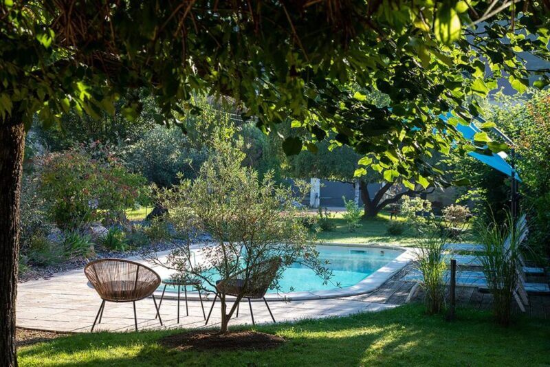 Two wicker chairs and a small table sit on grass under a tree at O'myosotis, overlooking a rectangular swimming pool in a lush, sunlit Sarlat garden.