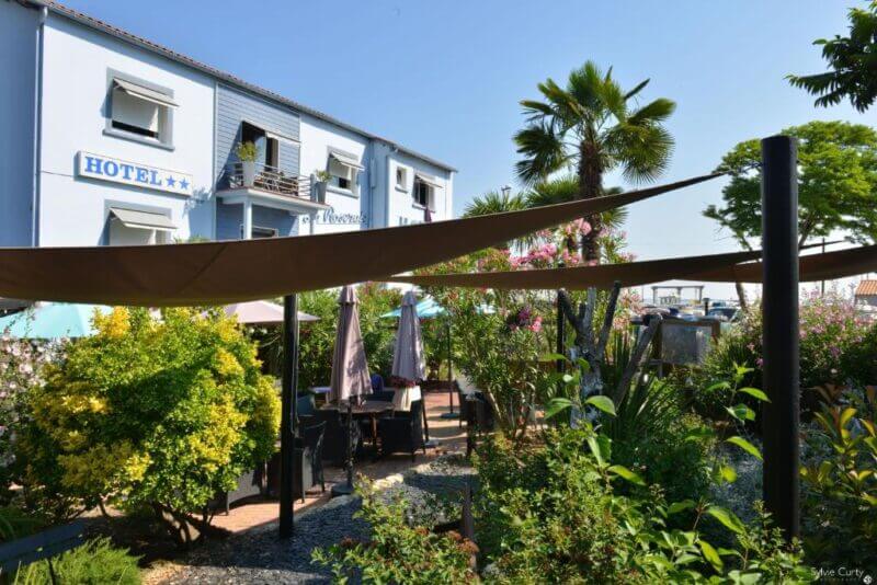 Outdoor seating area with tables and umbrellas surrounded by greenery in front of La Roseraie, a charming two-story Rochefort Hotel, basking in the sunshine.