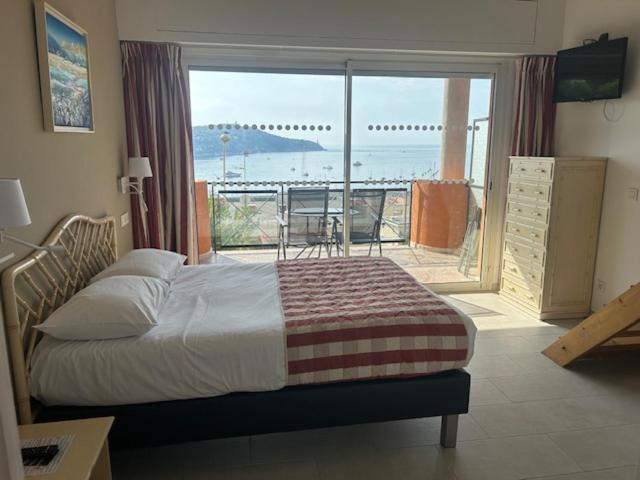 Hotel room at Hôtel La Flore in Villefranche-sur-mer with a double bed, dresser, wall-mounted TV, and sliding glass doors opening to a balcony with a sea view and outdoor table and chairs.