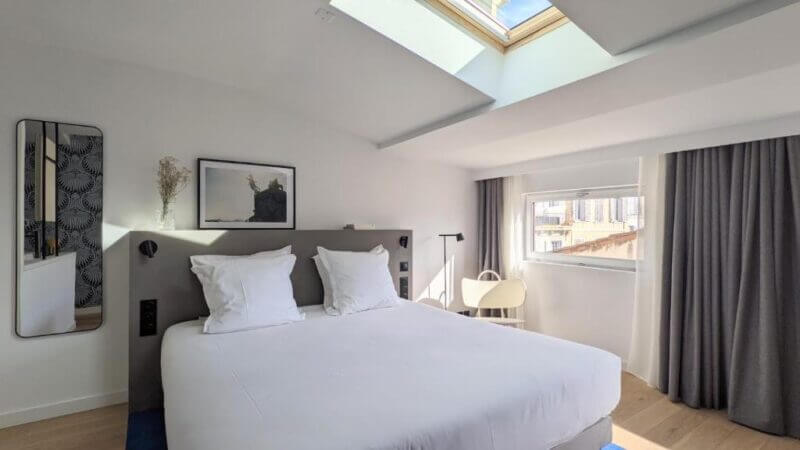 A modern hotel room at Maison Juste in Marseille, featuring a double bed with white bedding, wall art, a desk and chair by the window, and a skylight that fills the space with natural light.