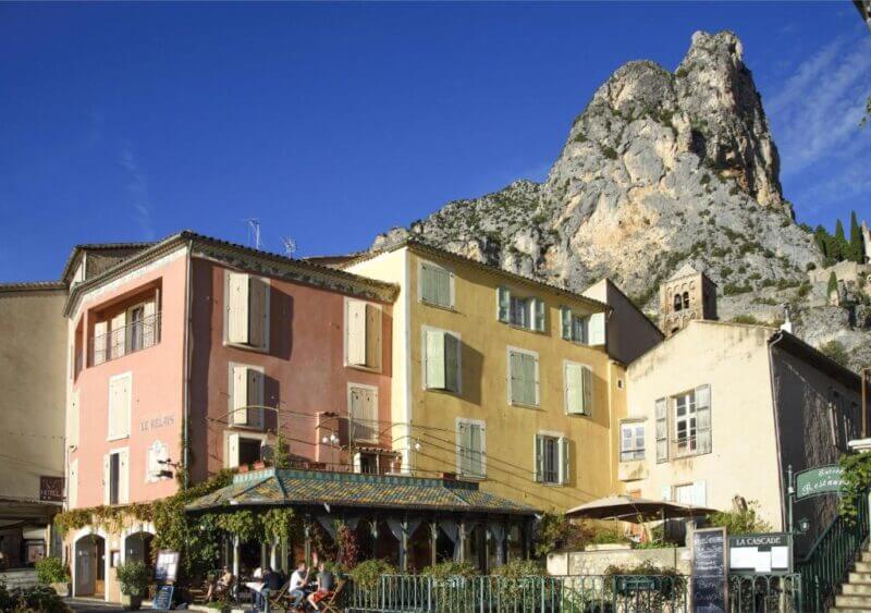 Colorful buildings with shuttered windows stand at the base of a rocky mountain near the Gorges du Verdon; people relax at an outdoor cafe, possibly Le Relais de Moustiers, under a clear blue sky.