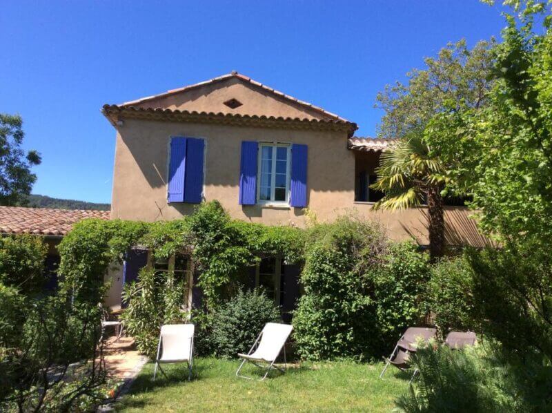Two-story house with tan walls and blue shutters, surrounded by green plants and trees, offers a peaceful retreat near Clos des Iris and Gorges du Verdon, with several lounge chairs on the grassy lawn.