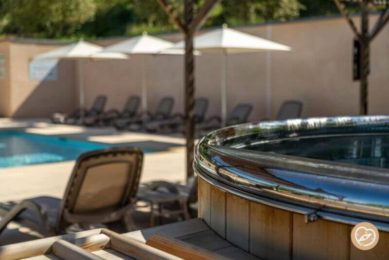 Close-up of a hot tub in the foreground at Les Restanques hotel, with a swimming pool, lounge chairs, and white umbrellas in the background on a sunny day near the stunning Gorges du Verdon.