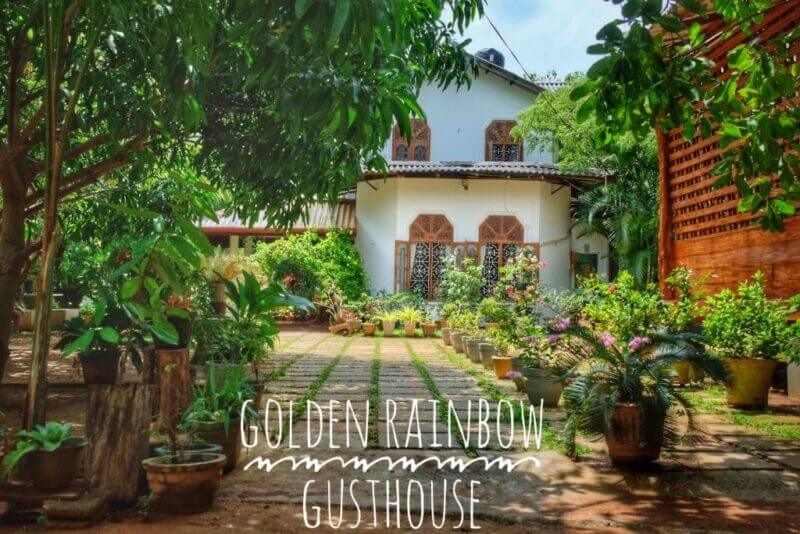 A white two-story guesthouse surrounded by lush greenery and potted plants, with Golden Rainbow Dambulla Guesthouse text at the bottom of the image.