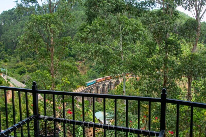 A train crosses the iconic 9 Arch viaduct bridge near Ella, winding through a lush forest and framed by a black metal balcony railing in the foreground, offering a stunning view.