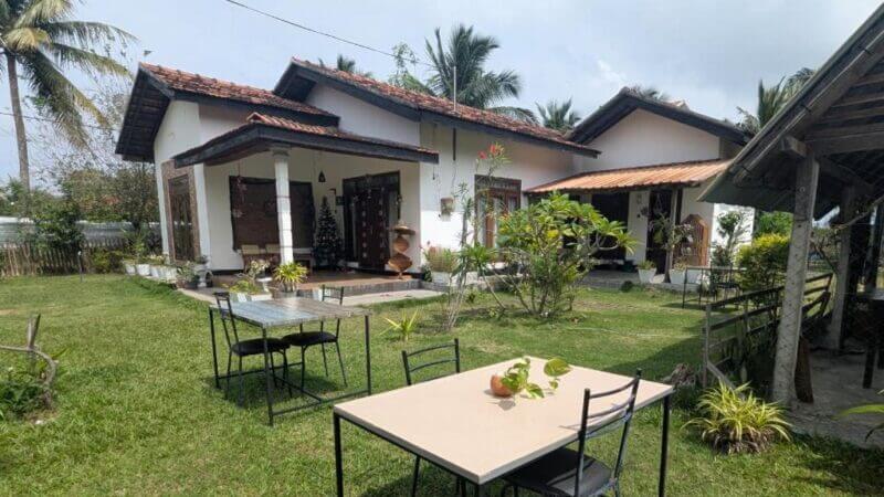 A single-story house with a tiled roof, surrounded by greenery and palm trees, Aiden Homestay in Pasikuda features an outdoor table and chairs set on the lawn for relaxing amidst nature.