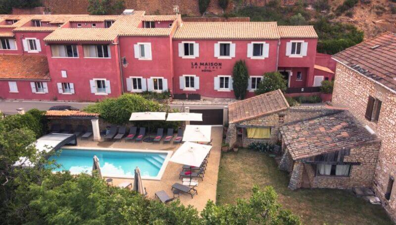 Aerial view of La Maison de Célou hotel in Rousillon, with a rectangular outdoor pool, lounge chairs, umbrellas, and rustic stone buildings in a landscaped courtyard reminiscent of Maison des Ocres.