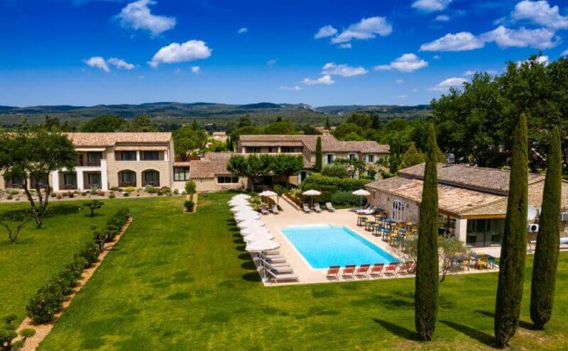 Aerial view of Mas de La Senancole villa complex in Gordes with a swimming pool, sun loungers, umbrellas, well-kept lawns, and surrounding trees under a blue sky with scattered clouds.