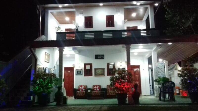 Two-story house at night with a covered porch, red and green lights illuminating potted plants, and outdoor seating visible on the right side—an inviting scene reminiscent of Green Village Dambulla, a tranquil Dambulla hotel.