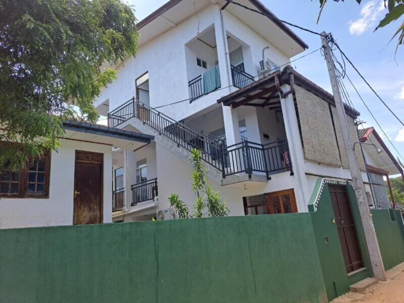 Two-story white building with balconies and exterior stairs, surrounded by a green wall and gated entrance, under a partly cloudy sky—welcome to B & B CHARM Dikwella.