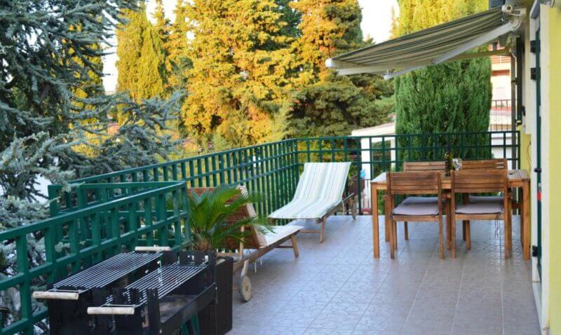 A spacious balcony in Vuković Apartments, Stari Grad, featuring a green railing, barbecue grill, lounge chair, wooden dining set for four, potted plants, and trees in the background.