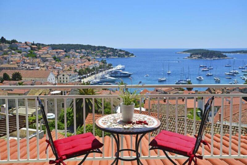 A small table with two red chairs on a balcony overlooks Hvar's marina and the sea, offering stunning views of the coastal town from these charming apartments under a clear blue sky.