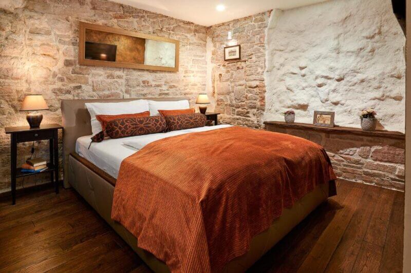 Cozy bedroom in Rustic Apartments Sibenik, featuring stone walls, a double bed with orange bedding, wooden flooring, and bedside tables with lamps.