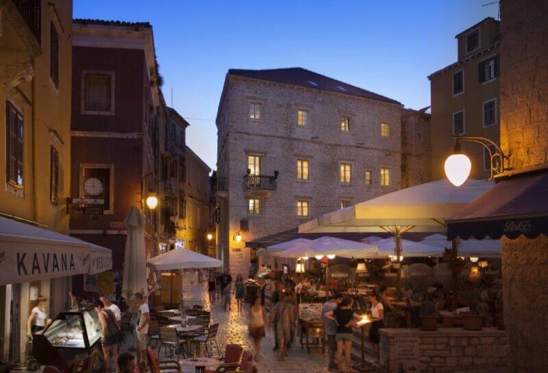 People walk through a cobblestone street lined with restaurants and outdoor seating in a historic European town at dusk, with warm lights illuminating the scene near Heritage Hotel Sibenik.