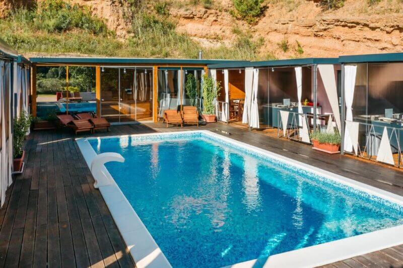 Rectangular outdoor swimming pool surrounded by wooden deck, lounge chairs, covered patios, and glass doors, set against a rocky Skradin hillside once part of ancient Illyria.