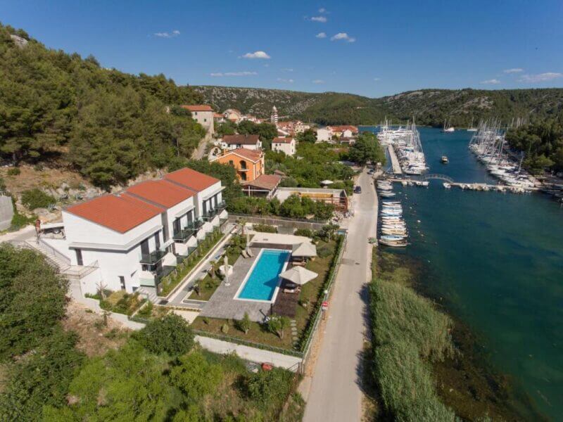 Aerial view of Hotel Bonaca Skradin’s modern white buildings with red roofs, a swimming pool, and gardens next to a marina with boats along a river or canal, surrounded by lush greenery and scenic hills.