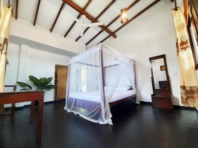 A spacious Sunstyle Mirissa bedroom featuring a four-poster bed draped in white mosquito netting, dark floors, wooden furniture, large windows, and a ceiling fan beneath a vaulted ceiling.