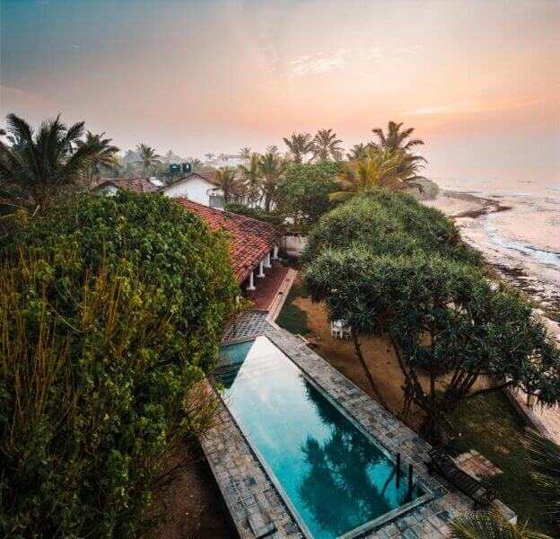 A narrow outdoor pool is surrounded by trees and tropical plants beside a red-roofed house at Saltasia Boutique, a charming boutique hotel near the Mirissa coastline at sunset.