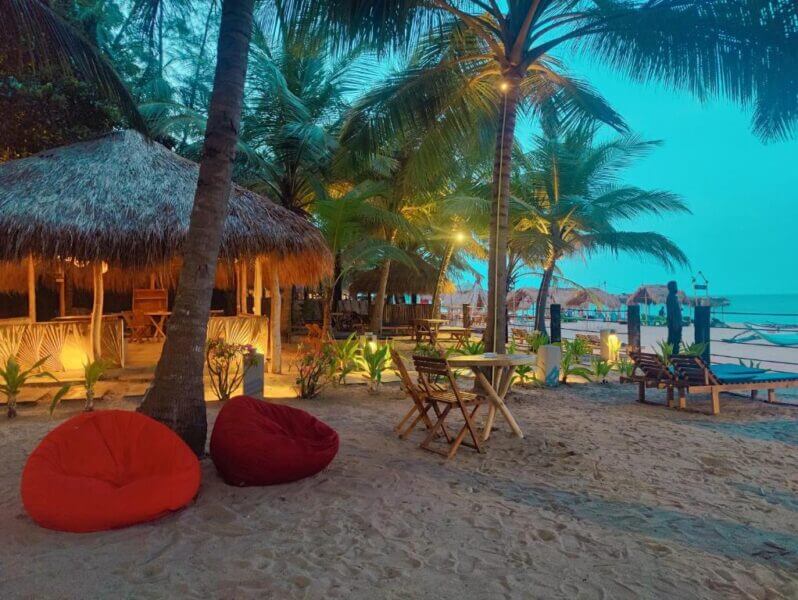 CHILL in style at Atteriya’s outdoor beachside seating area in Arugam, featuring wooden tables, chairs, red bean bags, palm trees, and thatched-roof huts just steps from the ocean at dusk.