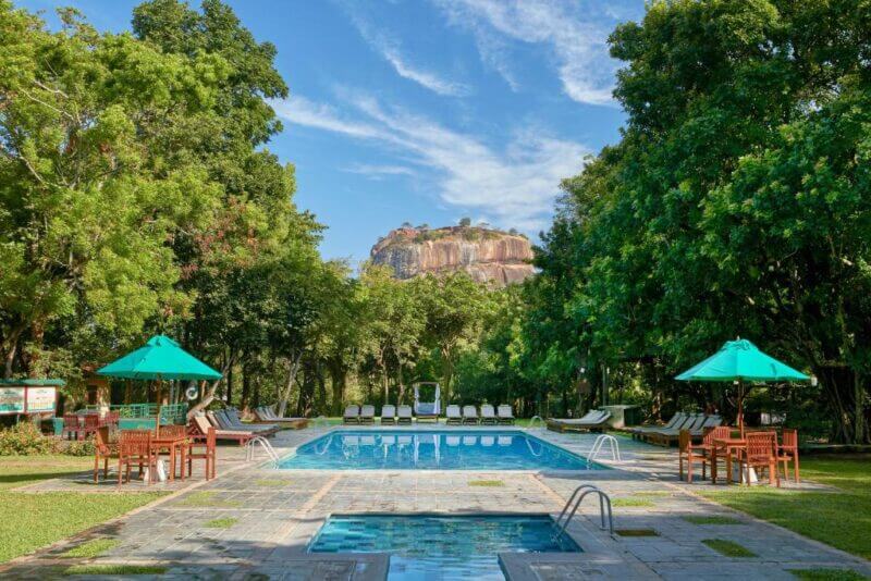 Rectangular swimming pool with lounge chairs and tables at Hotel Sigiriya, surrounded by trees, with a large rocky hill visible in the background under a blue sky.