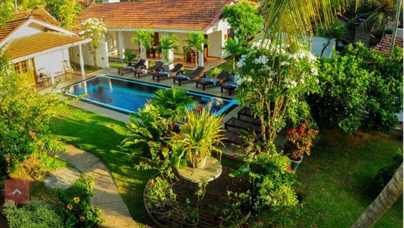 Aerial view of Sujeewani Villa in Negombo, featuring a tropical garden with a swimming pool, sun loungers, and two white buildings with red-tiled roofs, all surrounded by lush greenery.