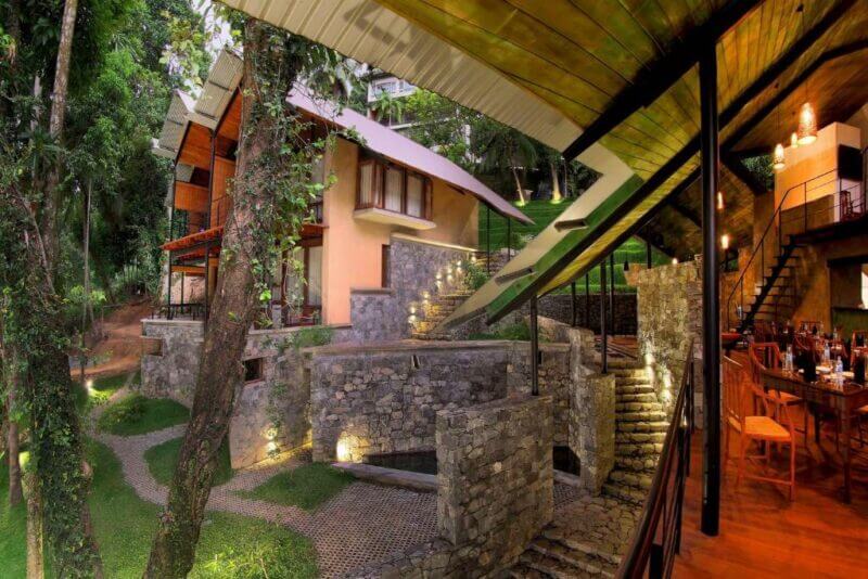 Modern stone and wood buildings surrounded by trees in Kandy, featuring outdoor stairs, pathways, and a dining area with tables and chairs—an inviting setting at SWP Eco Lodge.
