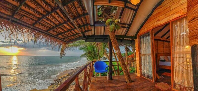 A wooden beach hut at Indika Beach Villa features an open veranda, a blue chair, and stunning ocean sunset views. Waves crash against rocks below, palm trees sway nearby—a tranquil Galle escape.