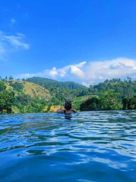 A person floats in a pool at View Cottage, gazing at green hills and the blue sky with scattered clouds in Ella.