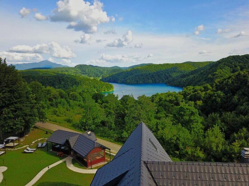 A scenic view of a lush green forest surrounding the blue Plitvice Lakes, with a few small buildings and Etno Garden outdoor seating areas in the foreground under a partly cloudy sky.