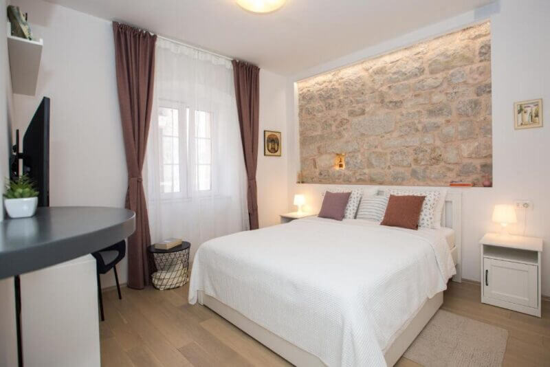 A modern bedroom at Liberty Rooms in Dubrovnik, featuring a double bed with white bedding, a stone accent wall, bedside tables with lamps, sheer-curtained windows, and a TV on a desk.