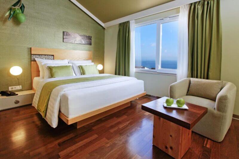 Modern Bol Boutique Hotel room featuring a double bed with green and white bedding, armchair, wooden table topped with green apples, and a large window offering a stunning ocean view.